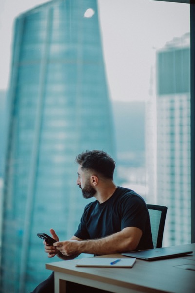 A man sitting at a desk by a high-rise window, phone in hand, gazing at the city skyline