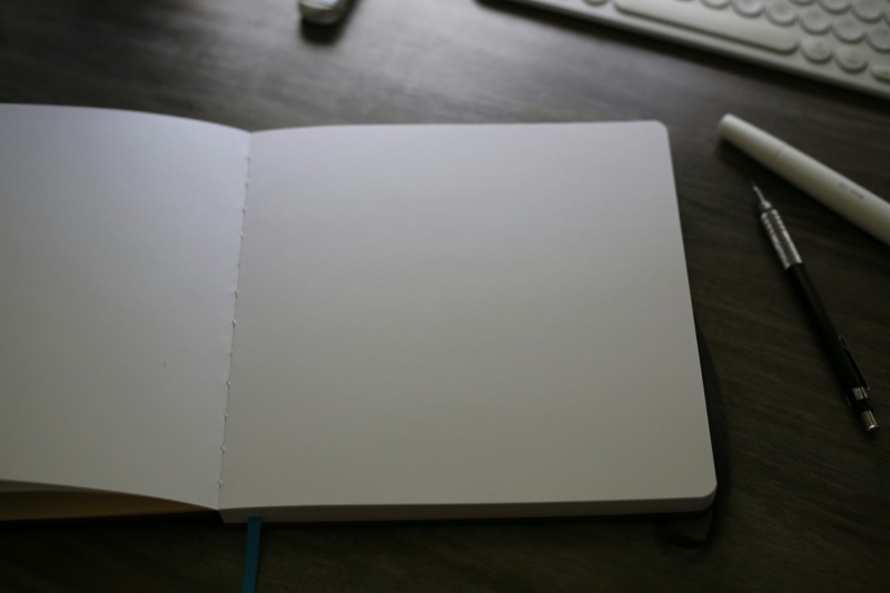 An open blank notebook on a dark wood desk next to a pen and keyboard