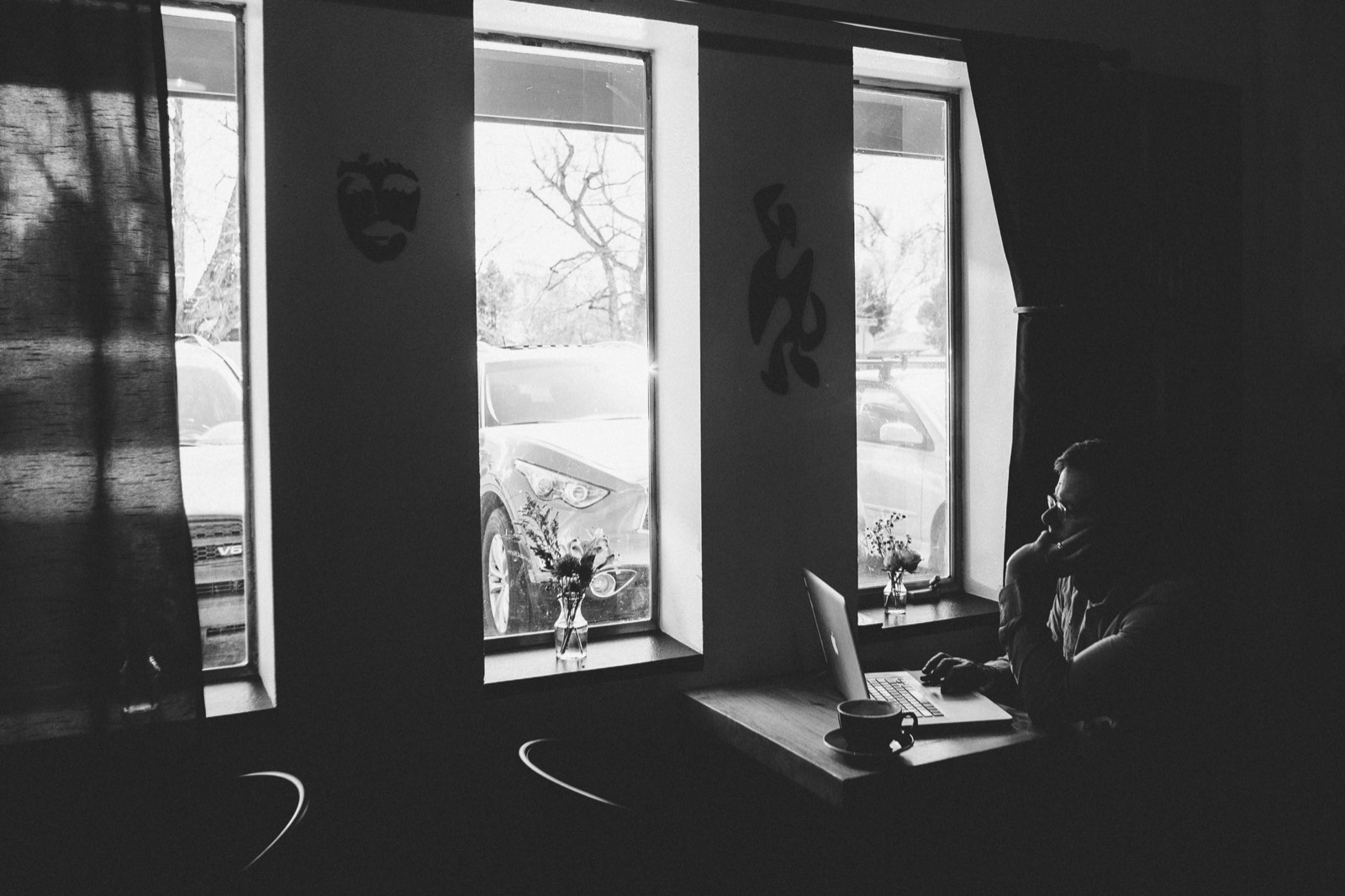 A person in silhouette at a window-side desk, laptop open, one hand resting on their chin, looking out
