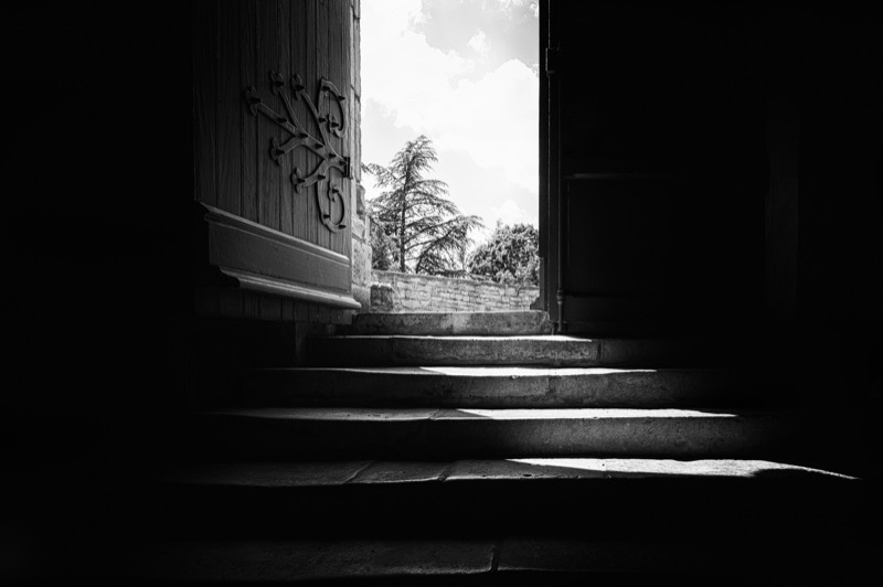 Black-and-white view through a heavy open doorway looking out to daylight and trees beyond