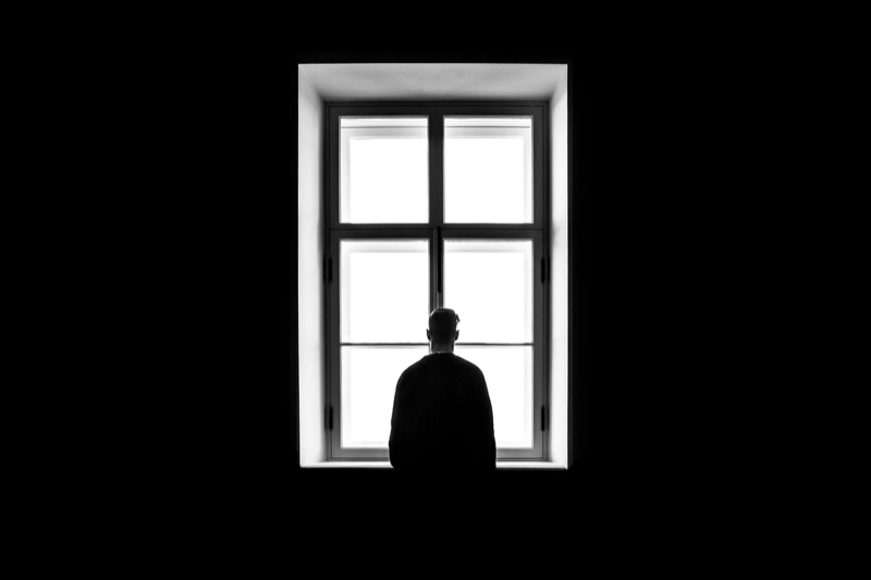 Silhouette of a person standing alone at a tall window in a dark room