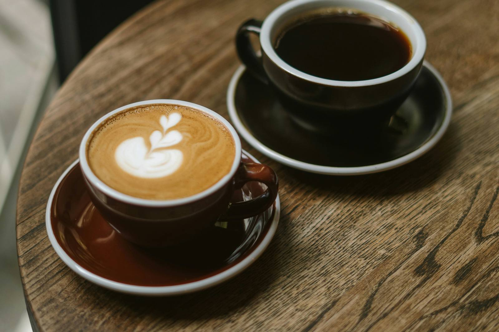 A latte and a cup of black coffee sitting side by side on a round wooden café table