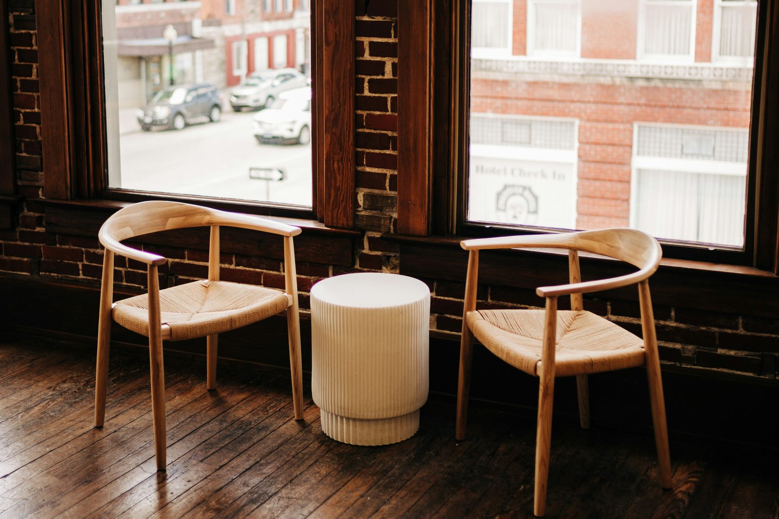 Two light wooden chairs facing each other across a small round table, in front of loft windows looking onto a brick street