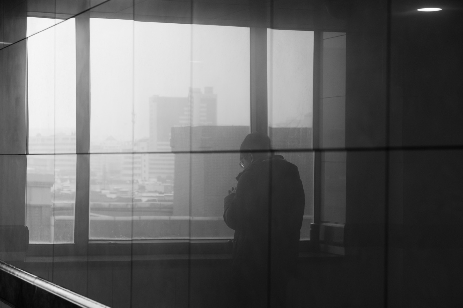 A person standing in silhouette at a tall window, looking out over a hazy city skyline