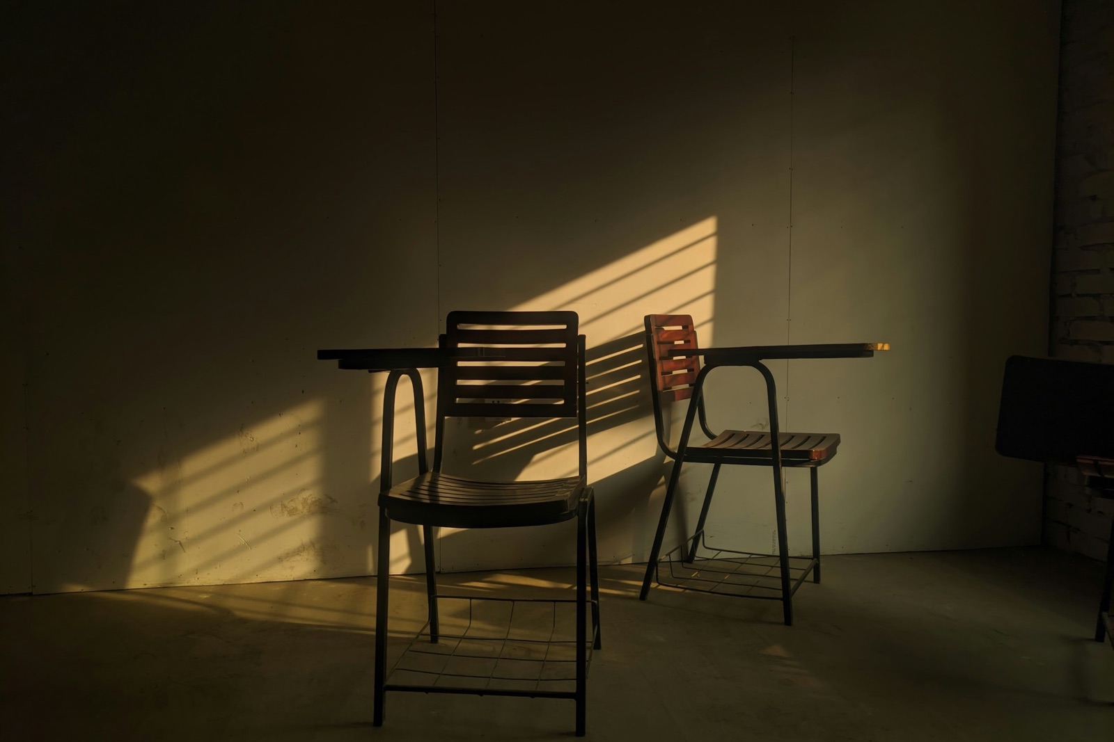 Two metal chairs at a plain table in a dim room, late sun falling in stripes across the wall