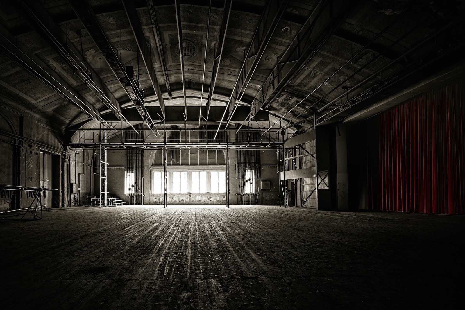 An empty hall with a worn wooden floor and a scaffold of stage rigging, lit by a bank of bright windows at the far end, with a red curtain hanging at the right edge