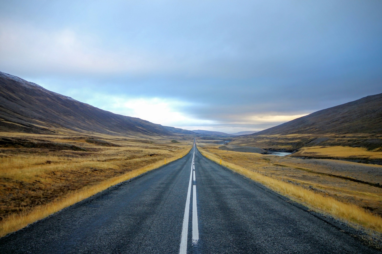 A two-lane road running straight through a wide valley of yellow autumn grass toward distant hills, under a low grey sky